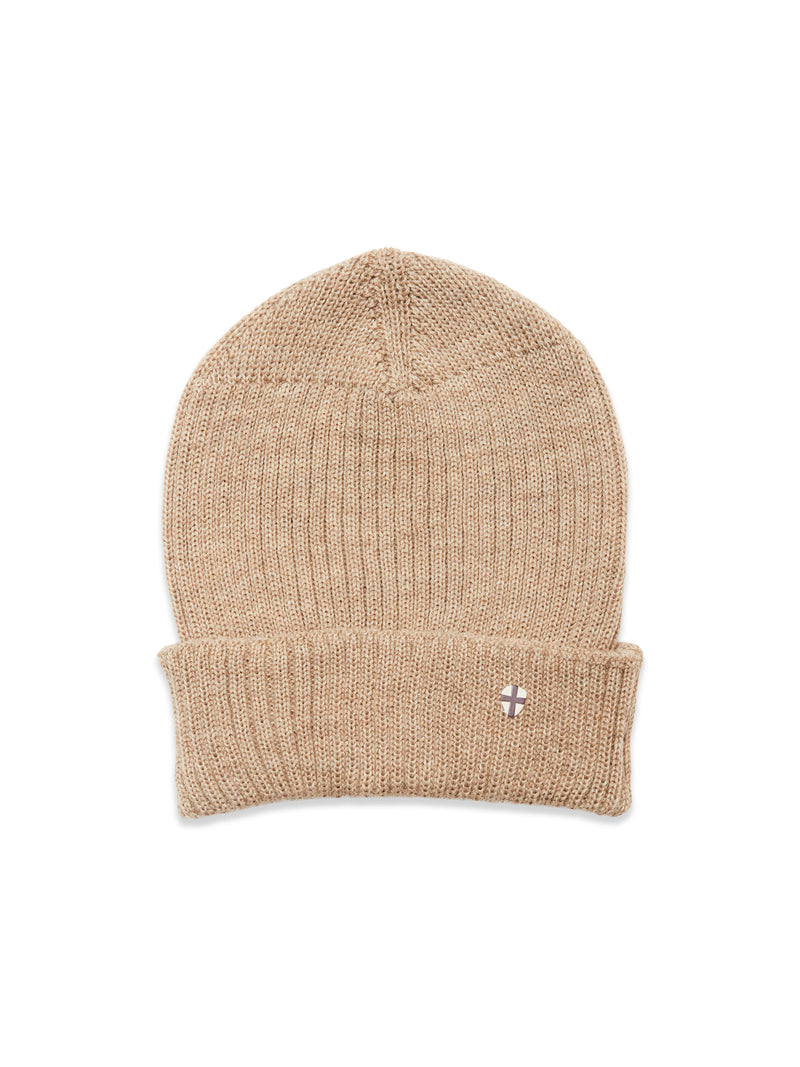 The Boshi Beanie - Oat by Blue de Gênes is a beige Merino wool rib beanie with a folded cuff and a small embroidered patch featuring a white and purple cross. Knitted in Denmark, its shown laid flat on a white background.