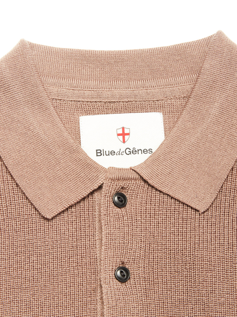 Close-up of the textured, light brown Porto Polo Knit - Fungi shirt by Blue de Gênes, featuring two black buttons and a collar label with the brand name and red cross emblem—a wool polo that exudes casual elegance.