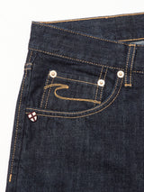 Close-up of the front pocket area of Marco Rinse Jeans by Blue de Gênes, in dark blue rinse wash 100% cotton denim with yellow stitching, metal rivets, and a shield-shaped enamel pin featuring a white cross on red.