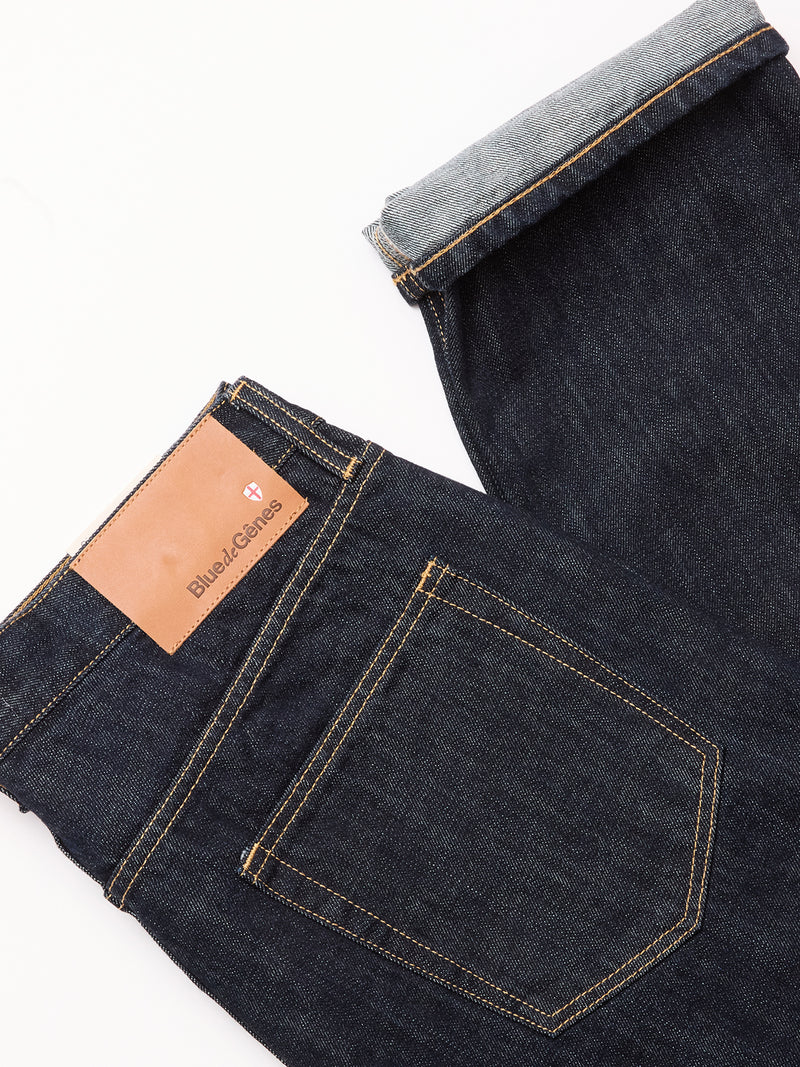 The Marco Rinse Jeans - Rinse Wash by Blue de Gênes are dark blue, relaxed-fit denim jeans with yellow stitching and a brown leather waistband patch; one cuff reveals the 100% cotton inner fabric.