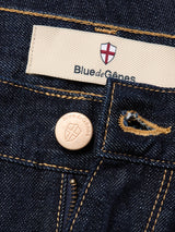 Close-up of Blue de Gênes Marco Rinse Jeans in a rinse wash, made from dark blue 100% cotton denim with a shield logo label, branded metal button, yellow stitching, and relaxed fit details on pockets and belt loops.