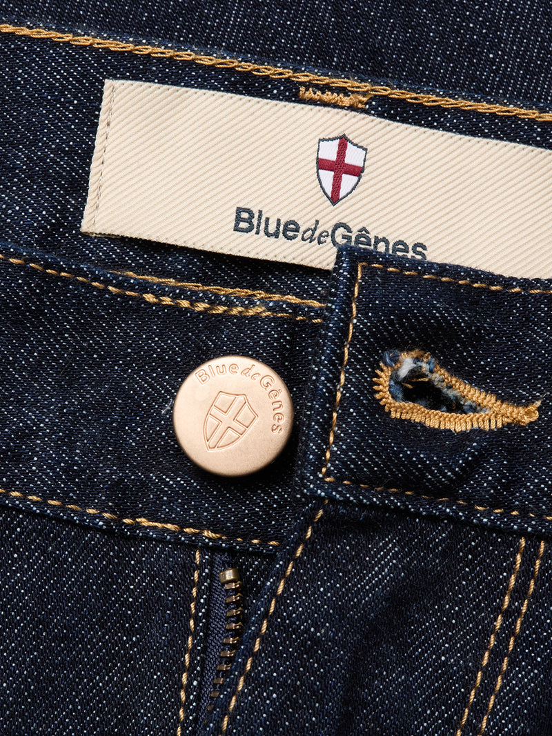 Close-up of Blue de Gênes Marco Rinse Jeans in a rinse wash, made from dark blue 100% cotton denim with a shield logo label, branded metal button, yellow stitching, and relaxed fit details on pockets and belt loops.