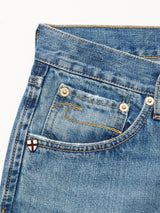 Close-up of blue denim jeans by Blue de Gênes, featuring a front pocket with gold stitching, a metal rivet, and a small red cross patch on white near the pocket. Made from 100% cotton denim. Product: Marco Used Jeans - Used.