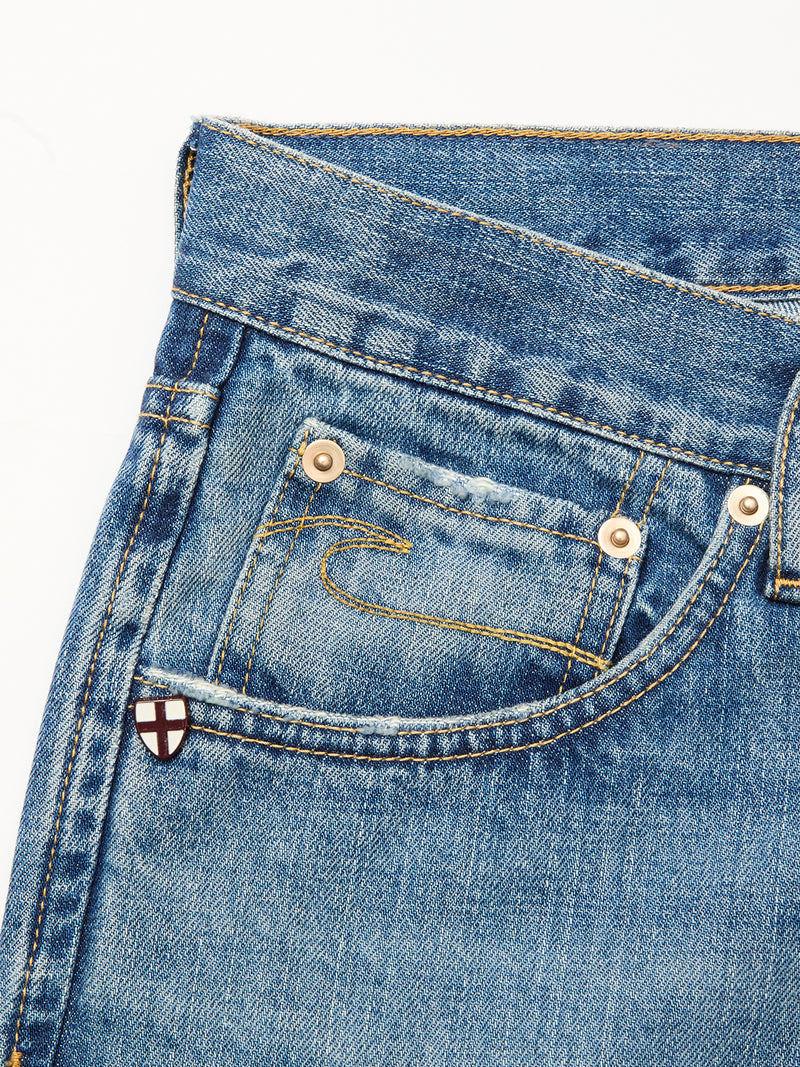 Close-up of blue denim jeans by Blue de Gênes, featuring a front pocket with gold stitching, a metal rivet, and a small red cross patch on white near the pocket. Made from 100% cotton denim. Product: Marco Used Jeans - Used.