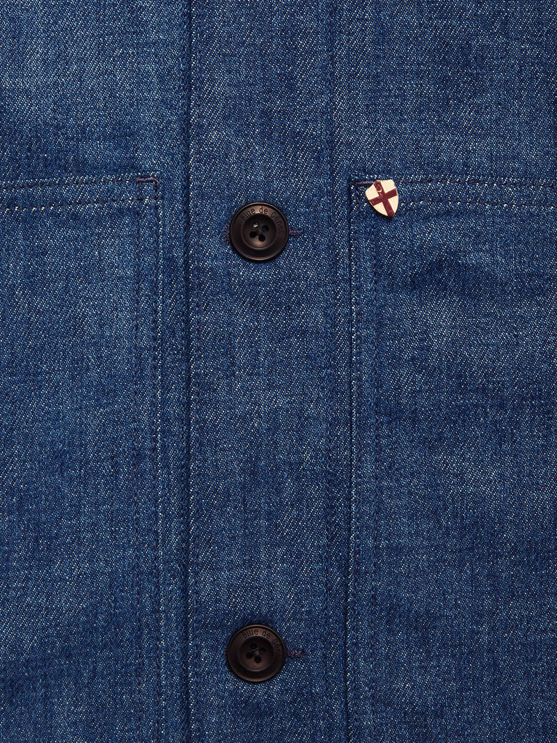 Close-up of the Blue de Gênes Yuma Fano Jacket in Denim Blue, a heavy cotton blend with two black buttons, two pockets, and a shield-shaped enamel pin featuring a red cross on white.