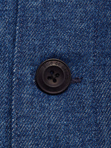 Close-up of a dark button engraved with Blue de Gênes on the Yuma Fano Jacket - Denim Blue, sewn onto blue denim fabric. Visible stitching and dark thread highlight the textured heavy cotton blend in this relaxed workwear-inspired piece.