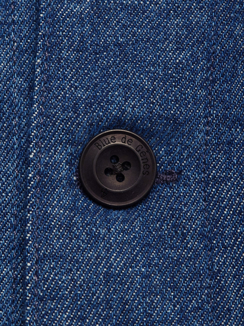 Close-up of a dark button engraved with Blue de Gênes on the Yuma Fano Jacket - Denim Blue, sewn onto blue denim fabric. Visible stitching and dark thread highlight the textured heavy cotton blend in this relaxed workwear-inspired piece.