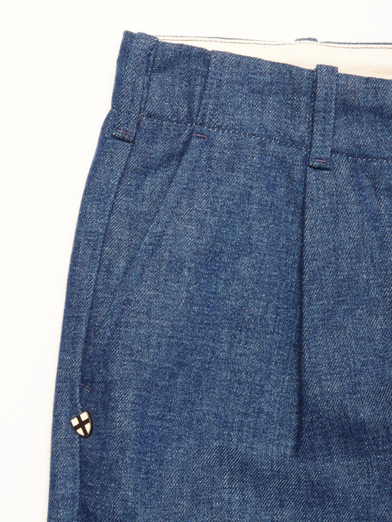 Close-up of Blue de Gênes Shogun Fano Pant in denim blue with pleats, waistband belt loops, and a small round pin—white with a black cross and red detail—near the pocket, crafted from heavy cotton blend.