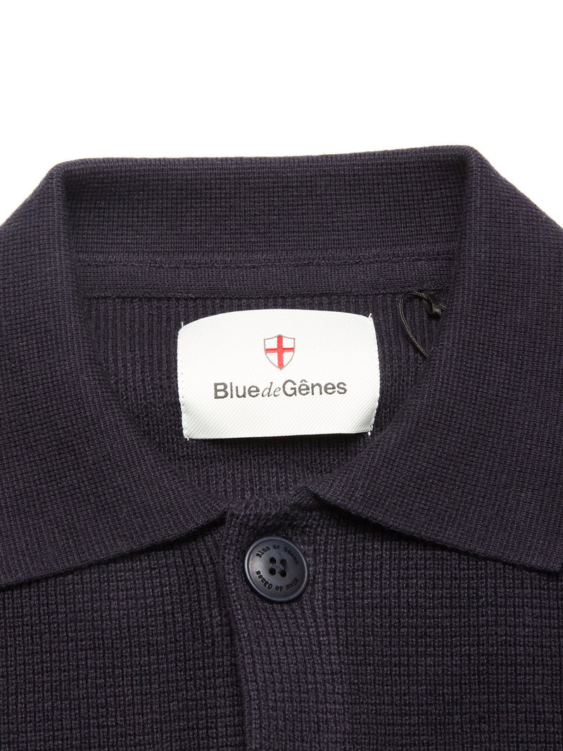 Close-up of a navy textured collared shirt with a black button and white Blue de Gênes label, perfect to pair with the Gifu Knit Cardigan - Navy from Blue de Gênes. A red shield logo sits above the text.