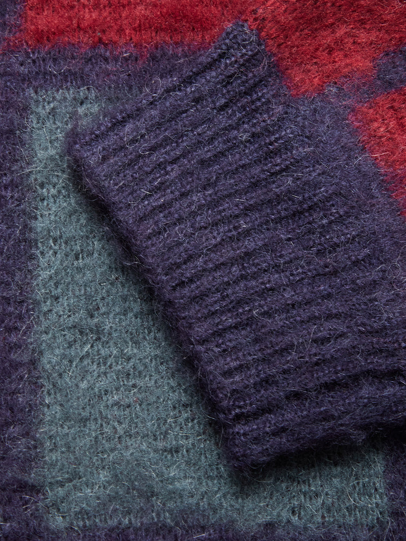 A close-up of a Block Knit Cardigan - Navy Pattern by Blue de Gênes, showing its textured dark purple knit sleeve on a multicolor background with red, green, and purple blocks.