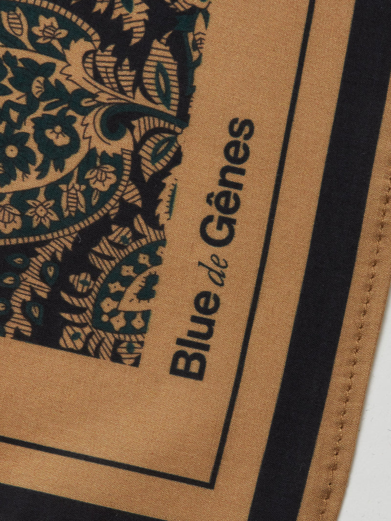 A close-up of the Siruku Scarf - Mixed Colours from Blue de Gênes shows an intricate black and teal floral pattern on tan, with the brand’s name printed in black along the edge of this stylish flower print scarf.