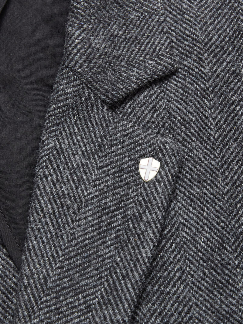 Close-up of the Fukui Blazer - Black Pattern by Blue de Gênes, a gray wool herringbone jacket with relaxed tailoring and a cross-design shield lapel pin on the left; its black inner lining is partially visible.