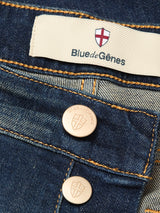 Close-up of Blue de Gênes Vinci Dark Vintage Jeans in mid blue denim, featuring a red and white shield logo label, two metallic branded buttons, comfort stretch fabric, and visible orange stitching.