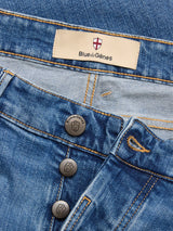 Close-up of BGvinci Hera Used Jeans by Blue de Gênes in blue-black denim with visible stitching, three metal shield-logo buttons, and a beige “Blue de Gênes” label above the waistband.