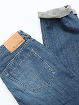 A pair of BGgiacomo Tim Jeans by Blue de Gênes in antique wash, featuring relaxed fit, visible back pocket stitching, a tan Blue de Gênes leather patch, and one cuffed leg revealing inner fabric with red and white stripe detail.