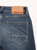 Close-up of the back of Blue de Gênes BGvinci Striker Jeans in dark distress, featuring a single back pocket and a brown leather patch with brand name and flag emblem on the waistband, made in Italy.