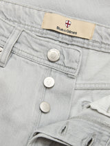 Close-up of Dolphin Grey BGgiacomo Color Jeans by Blue de Gênes, made from rigid light gray denim with silver buttons, waistband label, and an emblem with a red cross above the brand name.