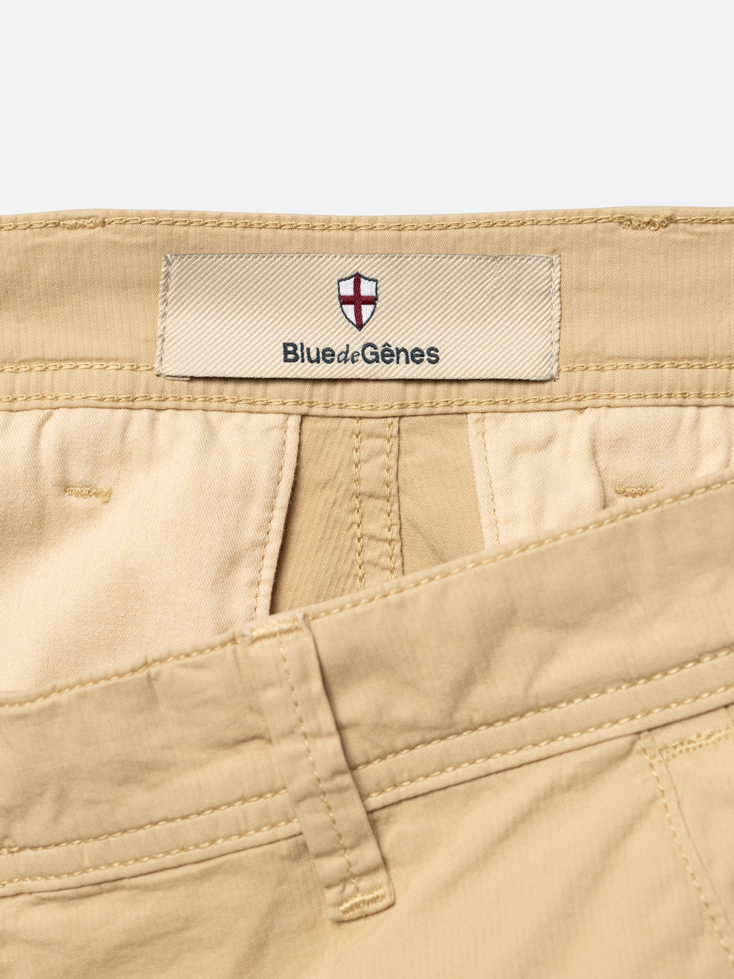 Close-up of Blue de Gênes BGmenzo Vecchio Chino in Pale Khaki, highlighting the waistband, belt loops, and inner fabric label with the brand and shield emblem—classic comfort stretch chinos with a vintage look.