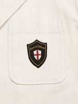 A close-up of the BGbaio Twill Blazer - Chalk by Blue de Gênes shows a white fabric pocket with a black and gold shield patch featuring "Blue de Gênes" and a red cross on a white background.