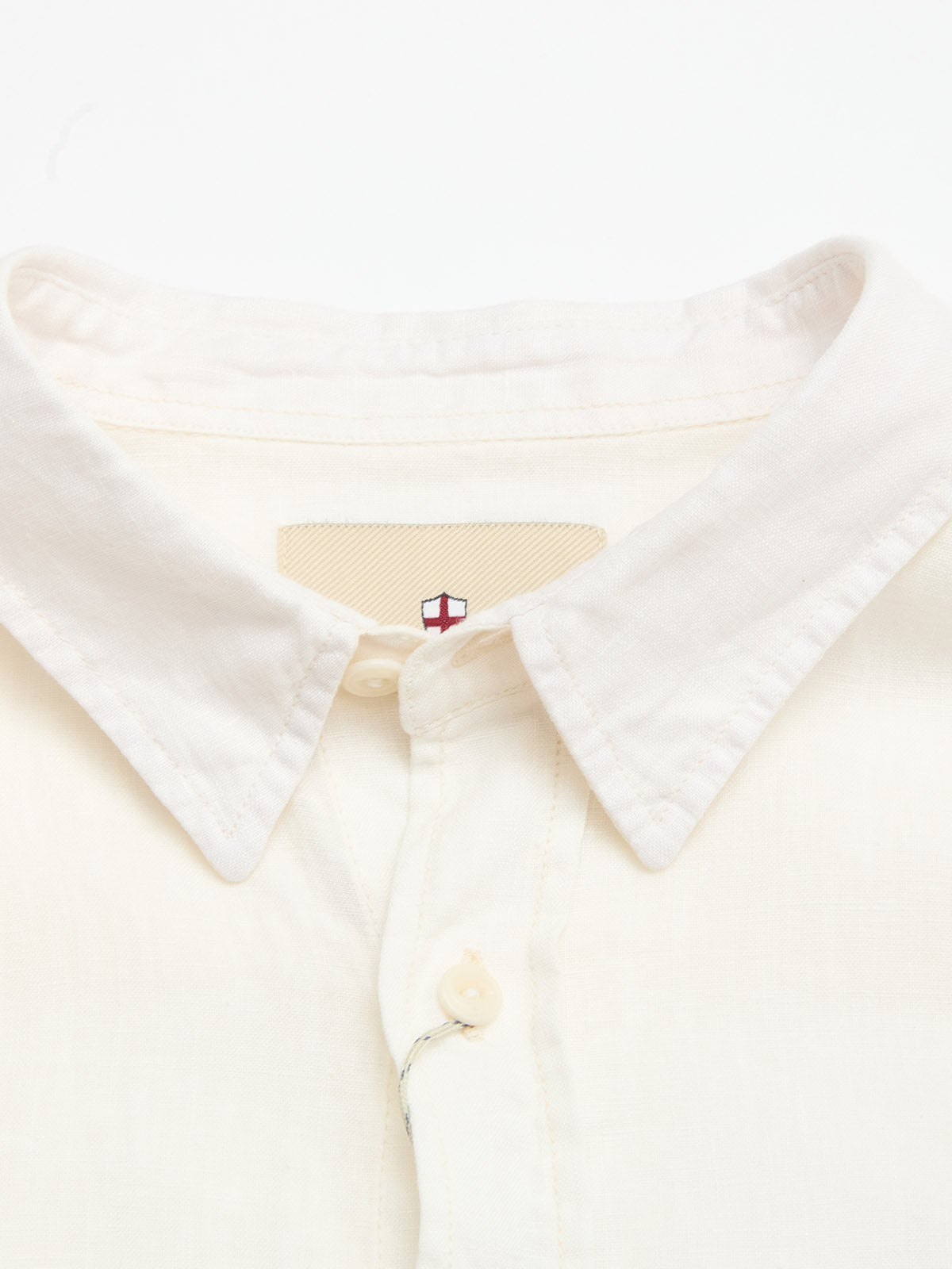 Close-up of the Blue de Gênes BGenric Hope L/S Shirt in Chalk. This off-white linen shirt features a cut-away collar, top button, and an inside tag with a red and blue cross logo. Neatly folded, it embodies breathable summer style.