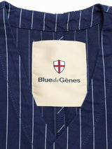Close-up of the BGlumen Toll Gilet - Navy Stripe by Blue de Gênes, featuring blue striped cotton and a cream label with the brand’s name below a red and white shield—highlighting its worker-inspired vest style.