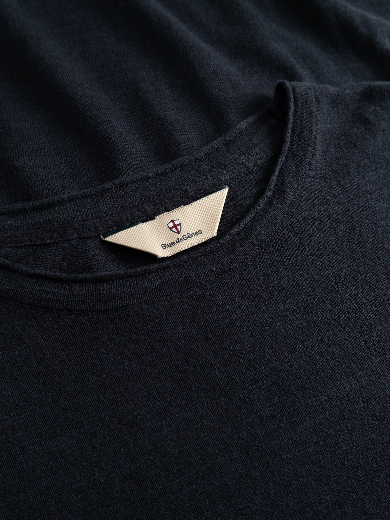 Close-up of a dark navy cotton-linen blend T-shirt, BGmonty Knit by Blue de Gênes, featuring a beige triangular neck tag with a small crest and partially visible branding. The soft fabric shows a subtle, textured finish.