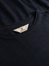 Close-up of a dark navy cotton-linen blend T-shirt, BGmonty Knit by Blue de Gênes, featuring a beige triangular neck tag with a small crest and partially visible branding. The soft fabric shows a subtle, textured finish.