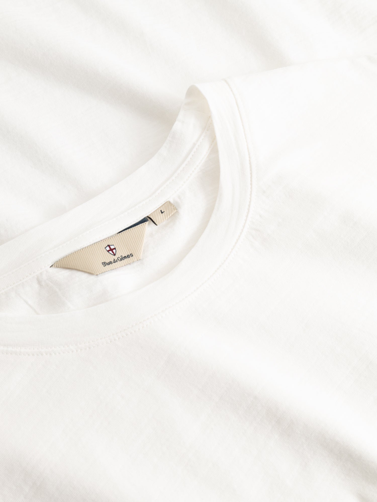 Close-up of the Blue de Gênes BGoscar T-Shirt - Snow Fall, neatly folded with a beige collar tag showing logo and text. Made from 100% cotton, this white crewneck blends comfort and style in every detail.