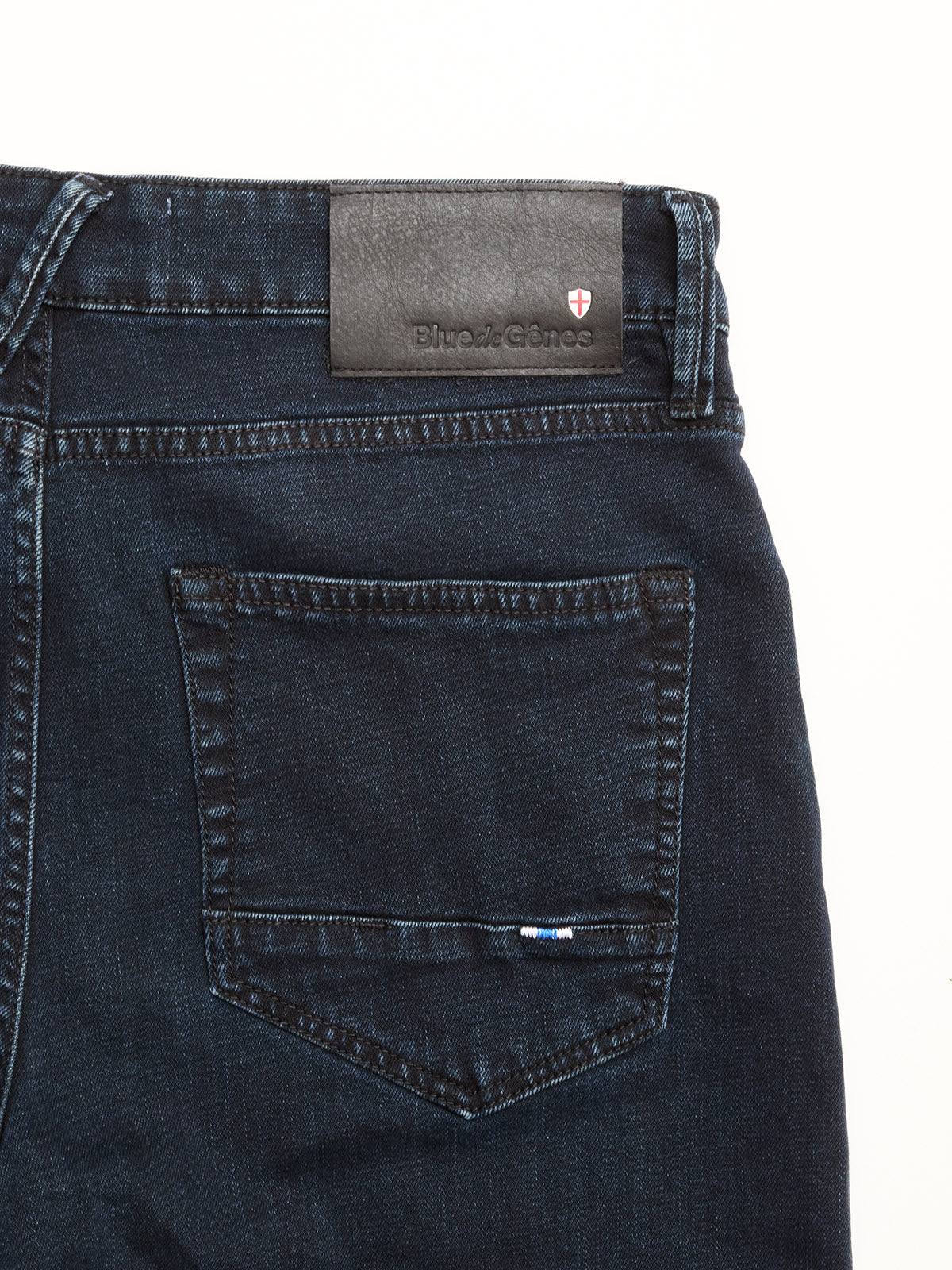 Close-up of the back pocket on BGvinci Black OD Jeans by Blue de Gênes, featuring visible stitching, a small label, and a black leather logo patch at the waist, crafted from blue-black stretch denim.
