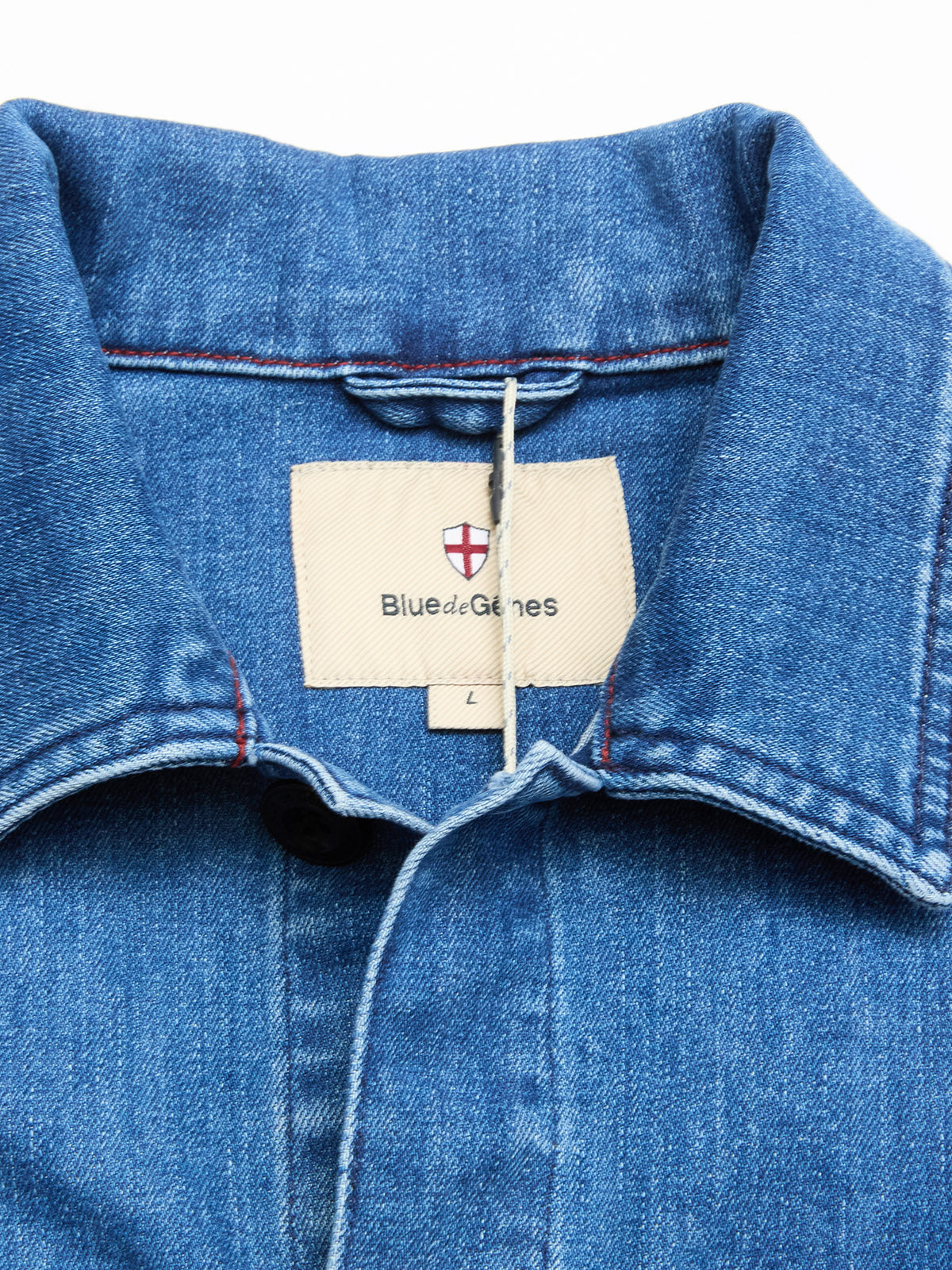 Close-up of a blue cotton blend BGpontus Denim Overshirt - Used by Blue de Gênes, showing the collar with an inner label featuring the brand name and a small red and white shield logo. A size L tag is visible below the label.