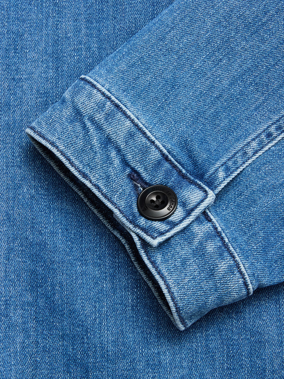 Close-up of the BGpontus Denim Overshirt - Used by Blue de Gênes, showing its blue cotton blend sleeve with a black button and detailed stitching, laid flat on matching denim.