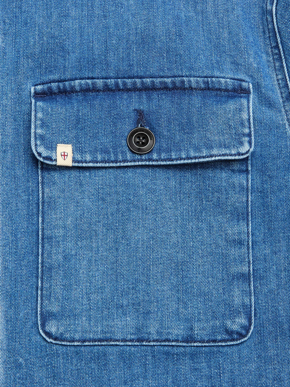 Close-up of a BGpontus Denim Overshirt - Used by Blue de Gênes, showing a blue cotton blend pocket with black button and a small white tag featuring a red shield logo on the upper flap. Relaxed fit and textured fabric are visible.