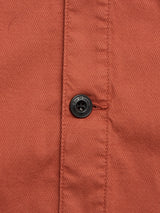 Close-up of the BGpontus Teo Overshirt in Copper Brown by Blue de Gênes, showing a rust-colored honeycomb cotton fabric with a black, engraved button sewn with orange thread near a vertical seam on this relaxed fit style.