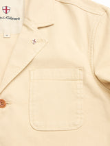 Close-up of the BGnorly Rodas Blazer in Pale Khaki by Blue de Gênes—a relaxed-fit cotton hemp blazer with a chest pocket, button, and an embroidered red cross shield logo on the collar. The Blue de Gênes label is visible inside.