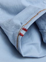 Close-up of the BGpaulo Pavia Pant in Blue Fog by Blue de Gênes, showing a light blue cotton stretch sleeve with brown edge stitching and two small red embroidered bars on slightly wrinkled fabric.