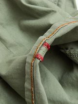 Close-up of BGpaulo Pavia Pant - Shadow Green by Blue de Gênes, featuring green cotton stretch fabric with an orange seam and a small red and white stitched label, highlighting detailed vintage military-inspired stitching and texture.