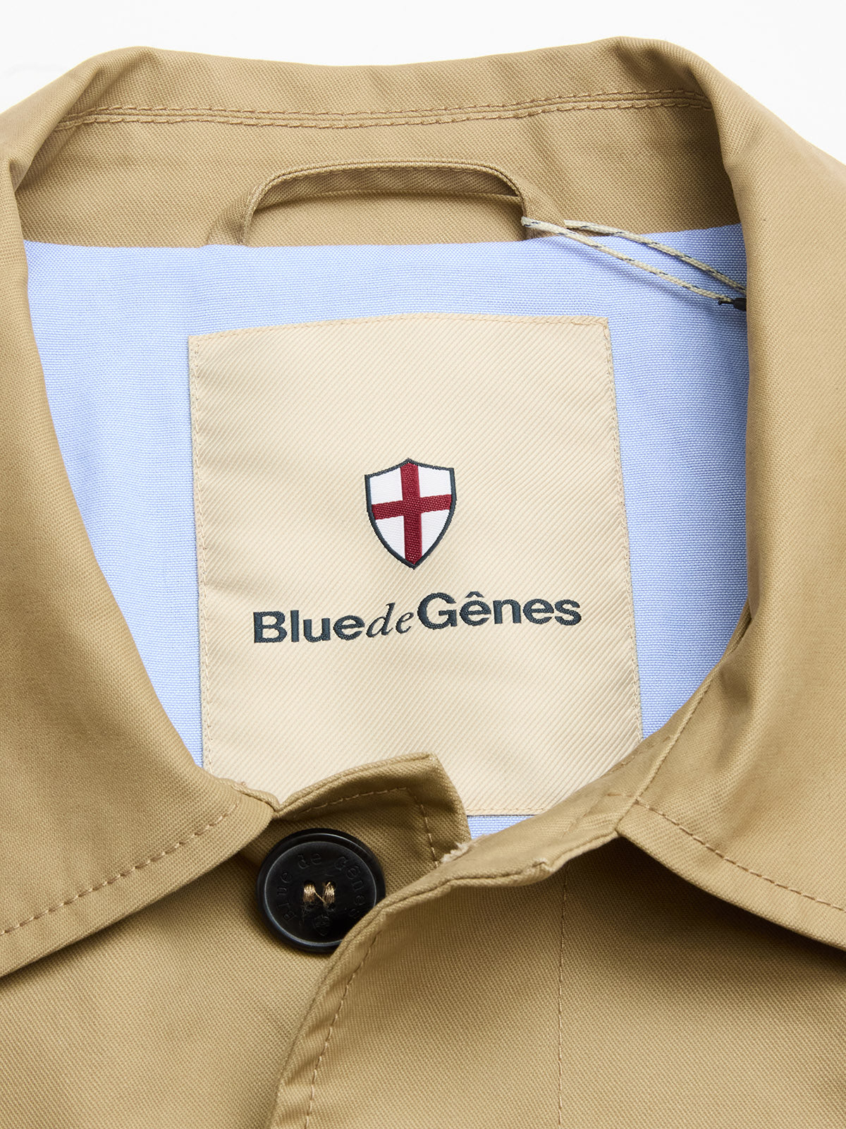 Close-up of the BGjacques Coat in pale khaki by Blue de Gênes, featuring a heavy cotton collar, shield logo with red cross, and a large Blue de Gênes label on light blue fabric with a classic black button for timeless workwear style.