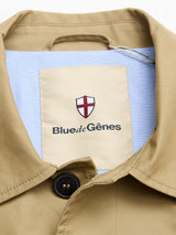 Close-up of the BGjacques Coat in pale khaki by Blue de Gênes, featuring a heavy cotton collar, shield logo with red cross, and a large Blue de Gênes label on light blue fabric with a classic black button for timeless workwear style.