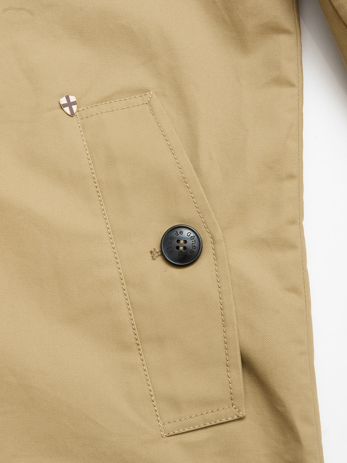 Close-up of the BGjacques Coat in Pale Khaki by Blue de Gênes, showing a heavy cotton pocket with a slanted flap, black button, and a shield-shaped pin featuring a white and brown cross above the pocket.
