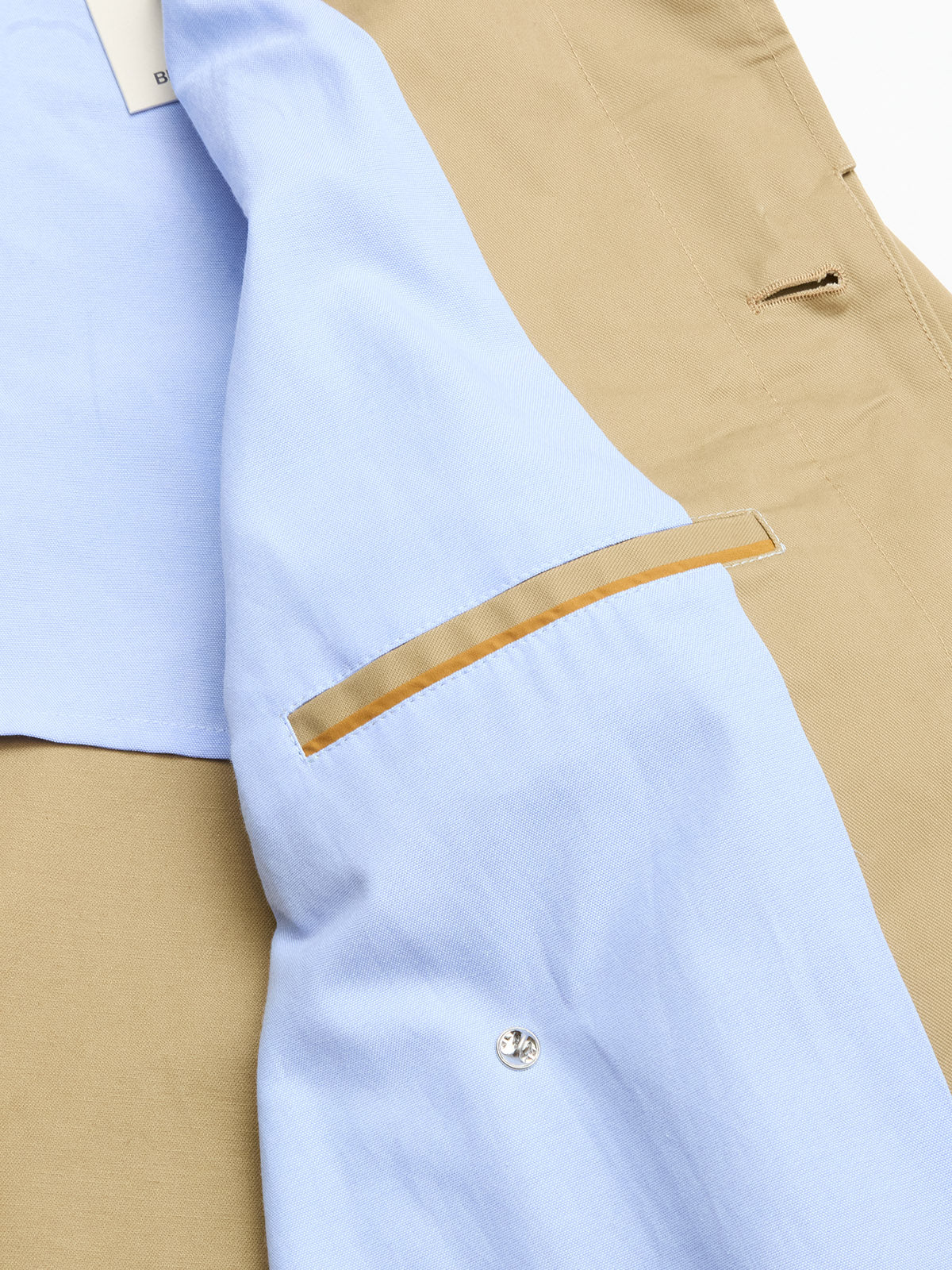 A close-up of the BGjacques Coat - Pale Khaki by Blue de Gênes highlights its beige heavy cotton fabric, light blue lining, interior pocket with brown trim, metal snap button, and a glimpse of a white tag.