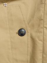 Close-up of a BGjacques Coat in Pale Khaki by Blue de Gênes, featuring heavy cotton fabric, visible stitching, a dark round button, and a small metallic shield emblem—evoking classic workwear style.