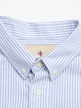 A close-up of the Blue de Gênes BGsander Oxford Shirt - Blue Stripe shows its button-down collar with the top button fastened and a small shield-shaped logo with a red cross inside the collar.