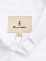 A close-up of the BGsander Oxford Shirt - White by Blue de Gênes shows its button-down collar and a beige label with a red and white shield logo, plus "Blue de Gênes" text. Made from 100% cotton, size L is marked below the label.