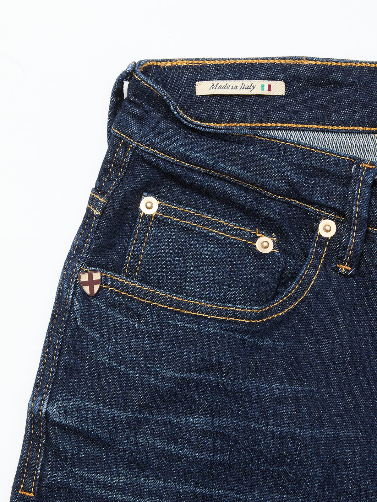 A close-up of Blue de Gênes Vinci Pala Dark V2 Jeans in dark blue denim shows gold stitching, metal rivets, a small front pocket with a shield-shaped pin, and a Made in Italy label above the waistband.