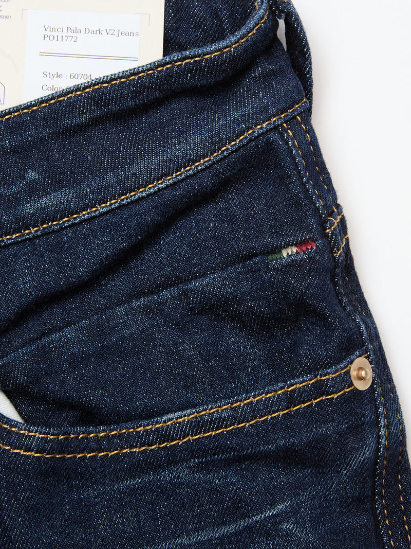 A close-up of Blue de Gênes Vinci Pala Dark V2 Jeans in dark blue stretch denim with yellow stitching, a brass rivet on the pocket, an Italian flag tag, and a partially visible clothing label at the waistband.
