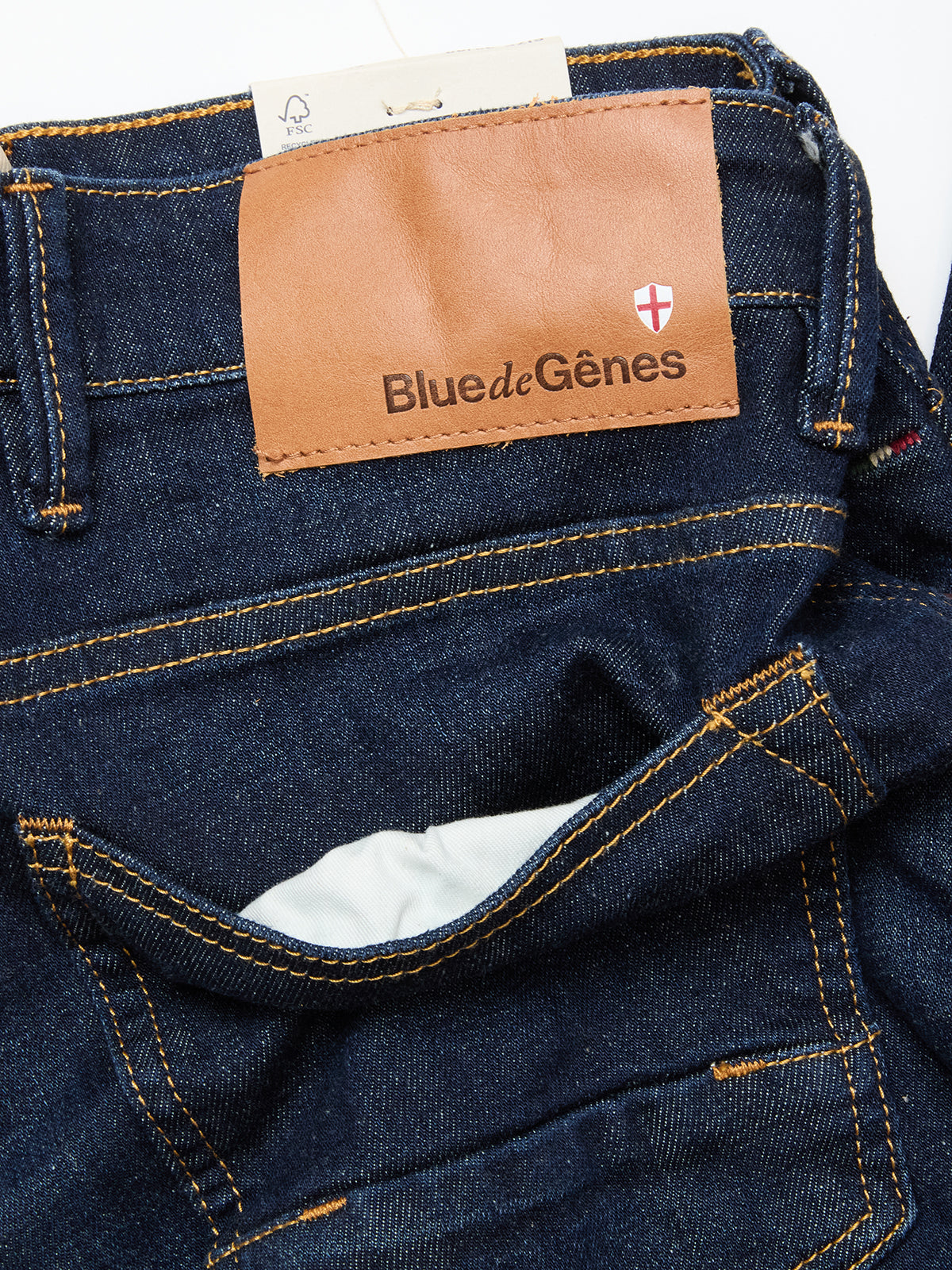 Close-up of the Vinci Pala Dark V2 Jeans by Blue de Gênes in dark blue denim, regular fit with orange stitching, a brown leather Blue de Gênes patch and shield emblem on the waistband, made from comfort stretch fabric for daily wear.