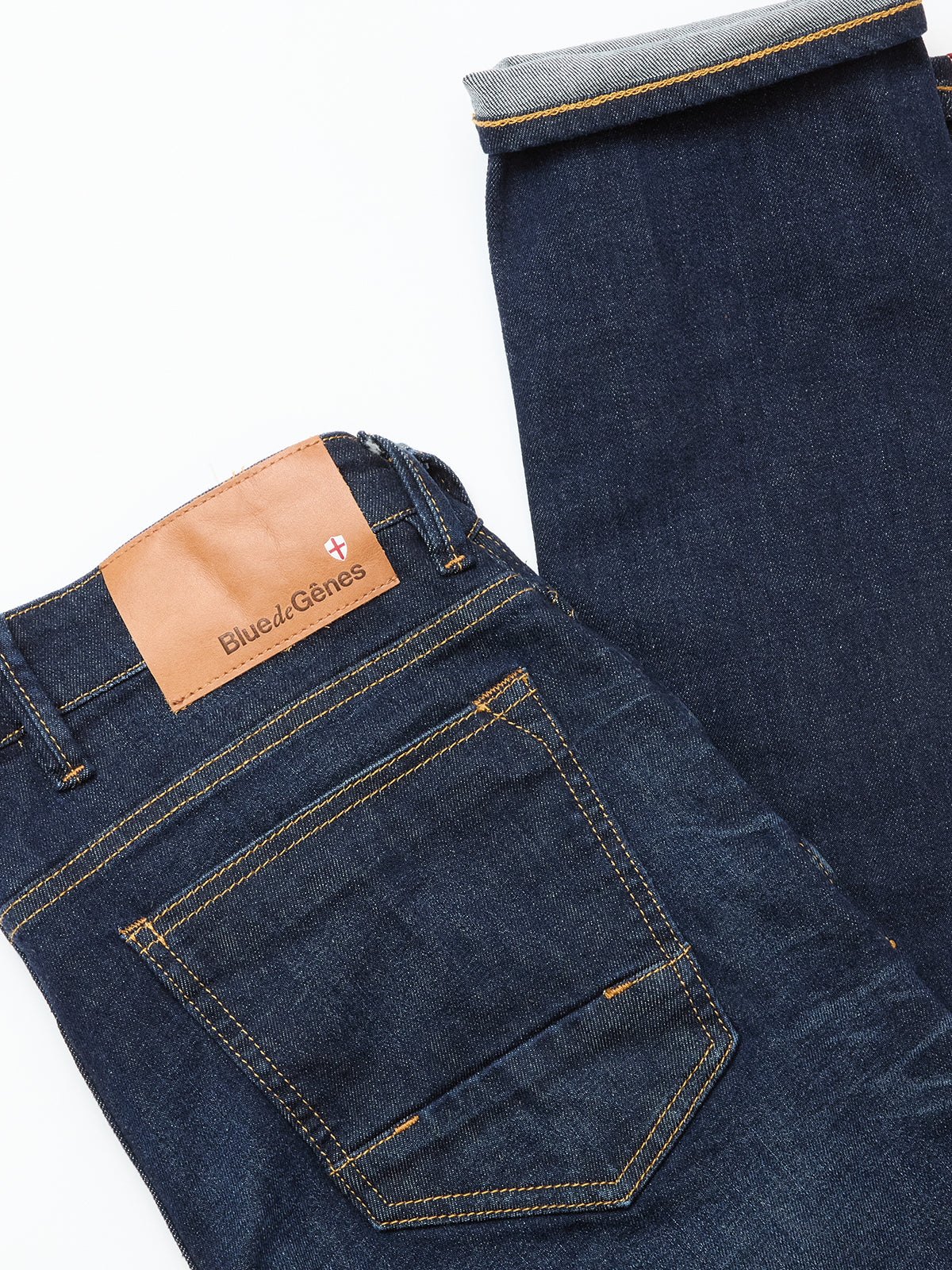 The Vinci Pala Dark V2 Jeans by Blue de Gênes are dark blue, regular fit denim with yellow stitching, a brown leather logo patch above the back pocket, and a turned-up leg to display the cuff and comfort stretch fabric.