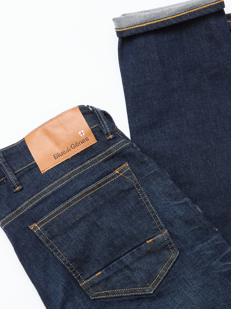 The Vinci Pala Dark V2 Jeans by Blue de Gênes are dark blue, regular fit denim with yellow stitching, a brown leather logo patch above the back pocket, and a turned-up leg to display the cuff and comfort stretch fabric.