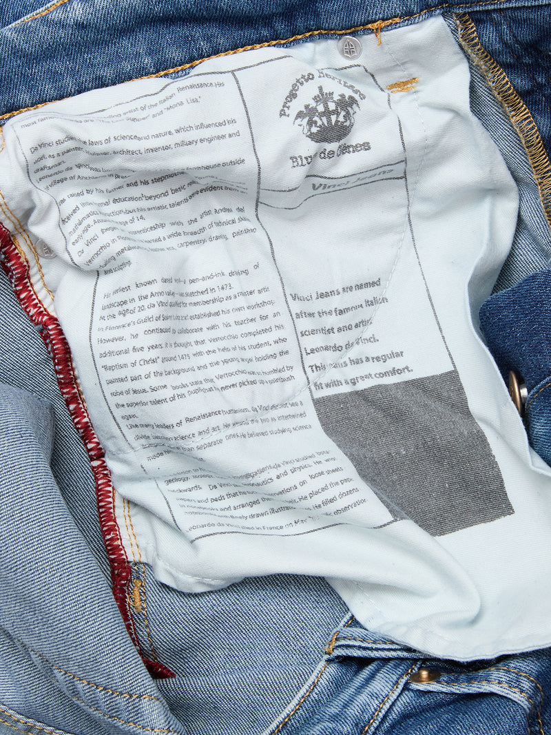 Close-up of the inside of Blue de Gênes Vinci Pala Used V2 Jeans in denim blue, showing a white label with black print and red stitching along the seam; text reveals regular fit, made in Italy, with some faded or obscured details.