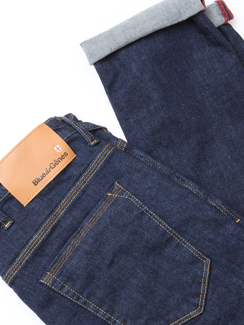 The Repi Dai V Dark V2 Jeans by Blue de Gênes are slim-fit, dark blue denim jeans with a brown leather patch. One cuffed leg reveals the lighter inner fabric and red seam stitching, highlighting the classic five-pocket design.